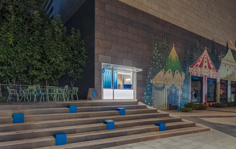 Illuminated kiosk between planted trees and decorative tent structures at evening