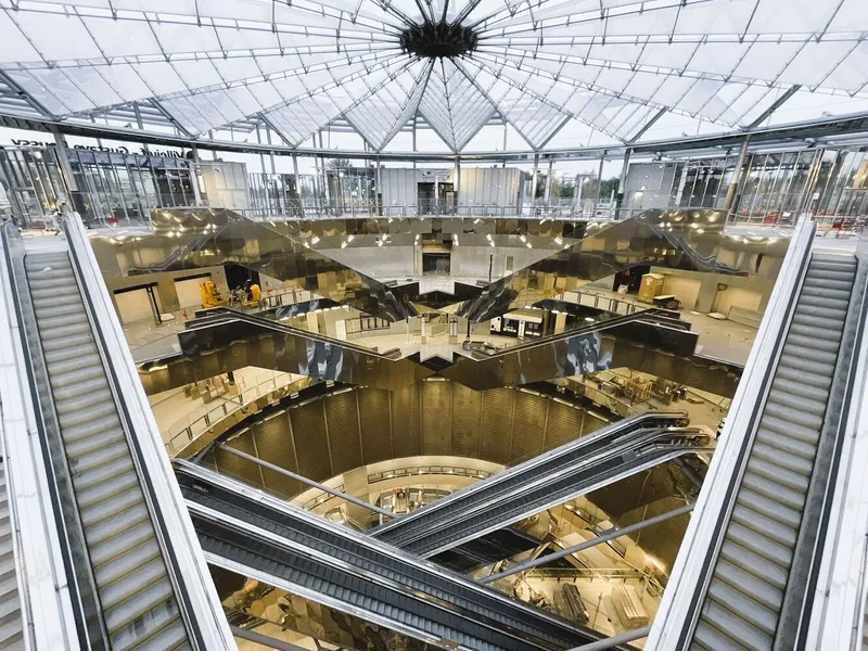 Central atrium with symmetrical escalators under a spoke-pattern glass roof and mirrored ceiling surfaces