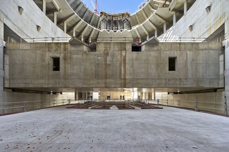 Interior view of circular concrete atrium with curved walls and incomplete roof structure under clear blue sky