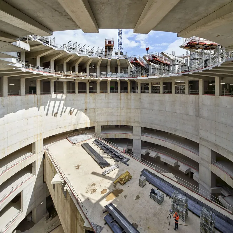 Multi-level circular concrete structure with exposed floor slabs, arched openings and construction scaffolding at roof level