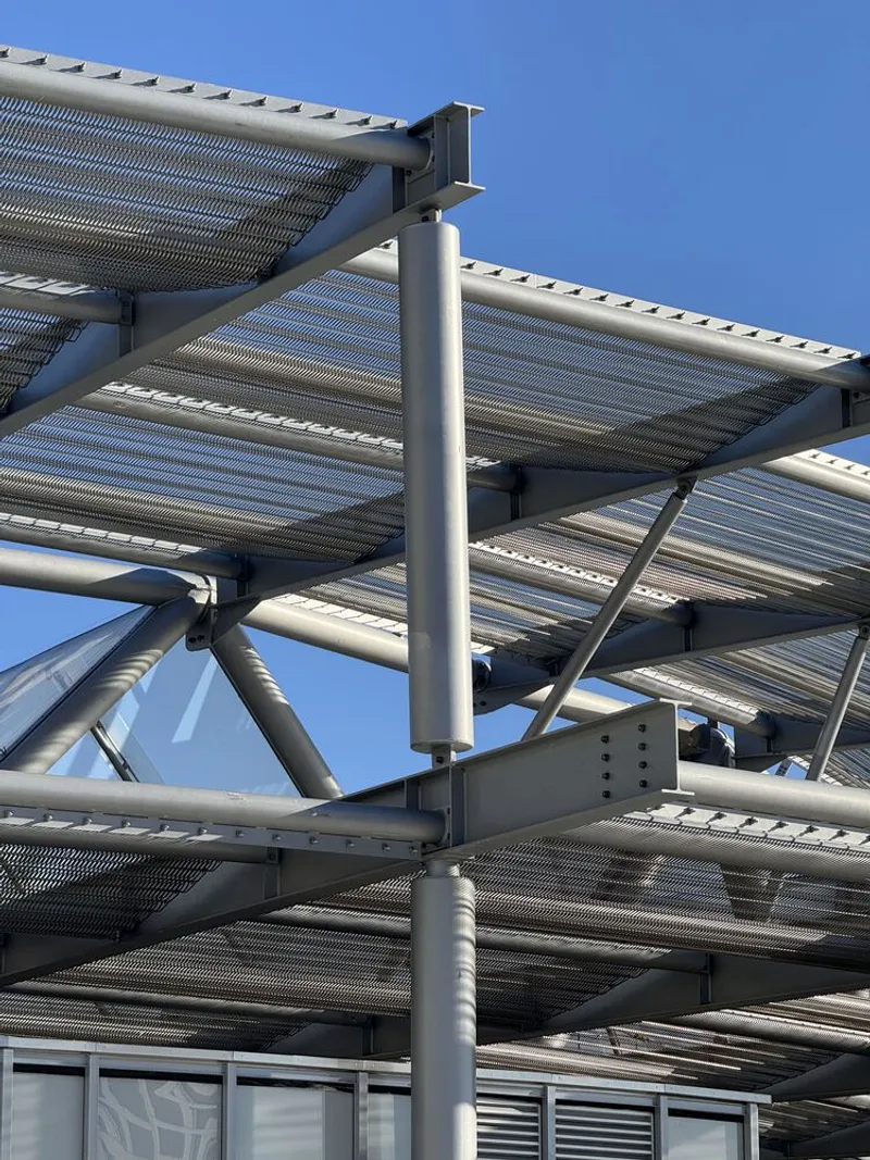 Detail of steel column connections and diagonal bracing beneath the perforated shade canopy against blue sky