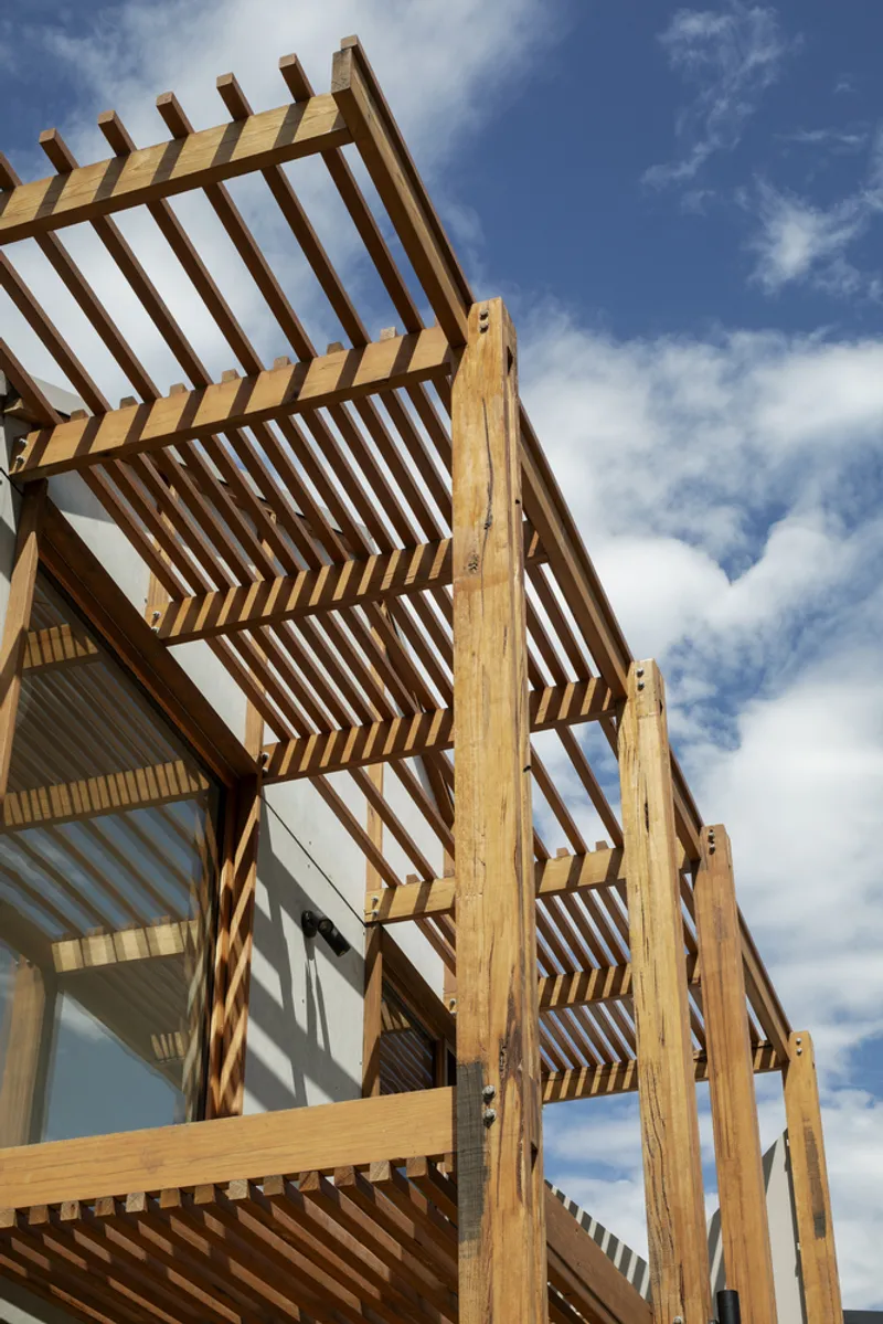 Upward view of the layered timber pergola screens creating shadow patterns against scattered clouds