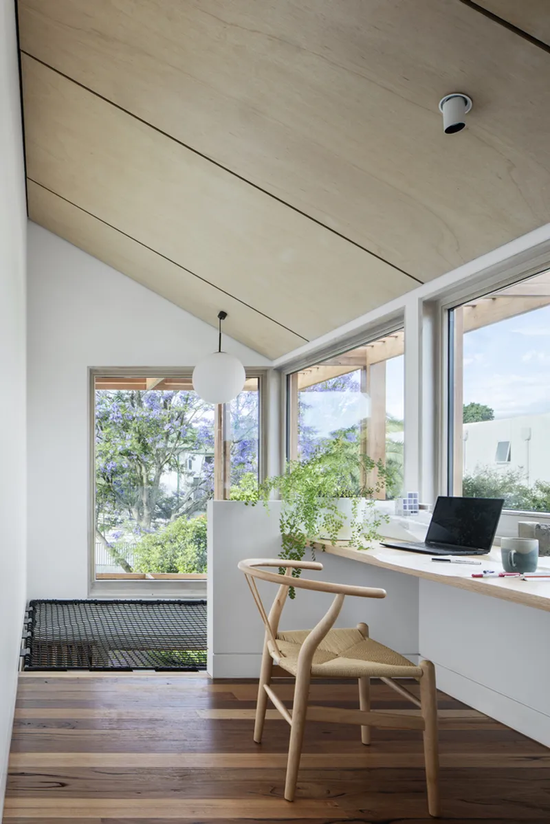 Home office with timber-lined ceiling and white desk overlooking a garden through corner windows