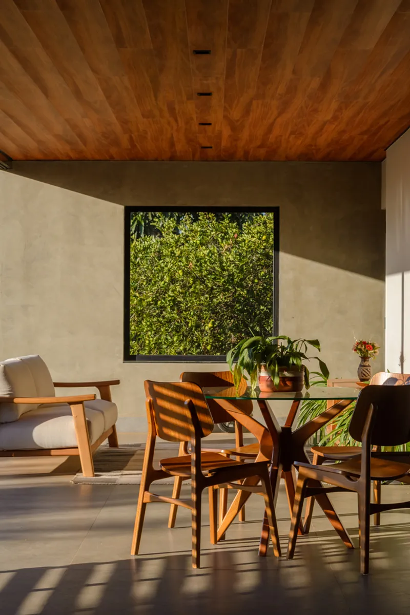 Dining area with timber ceiling, framed garden view, and sunlight casting shadows across the concrete floor
