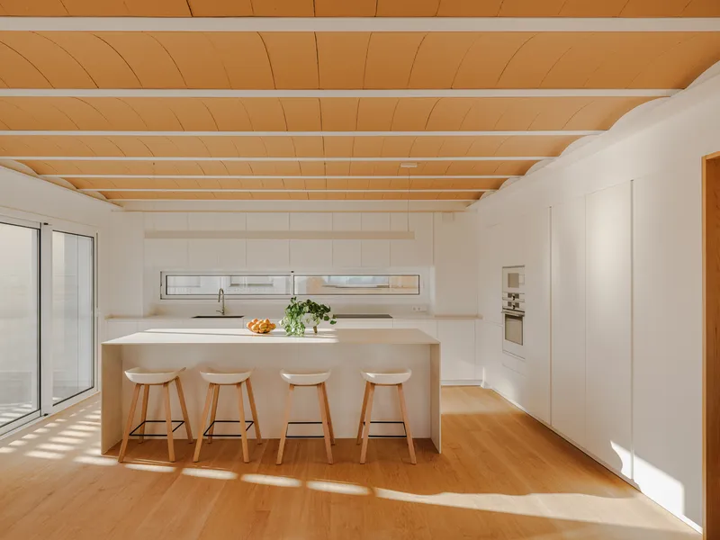 White kitchen island with timber bar stools beneath plywood ceiling with white exposed beams