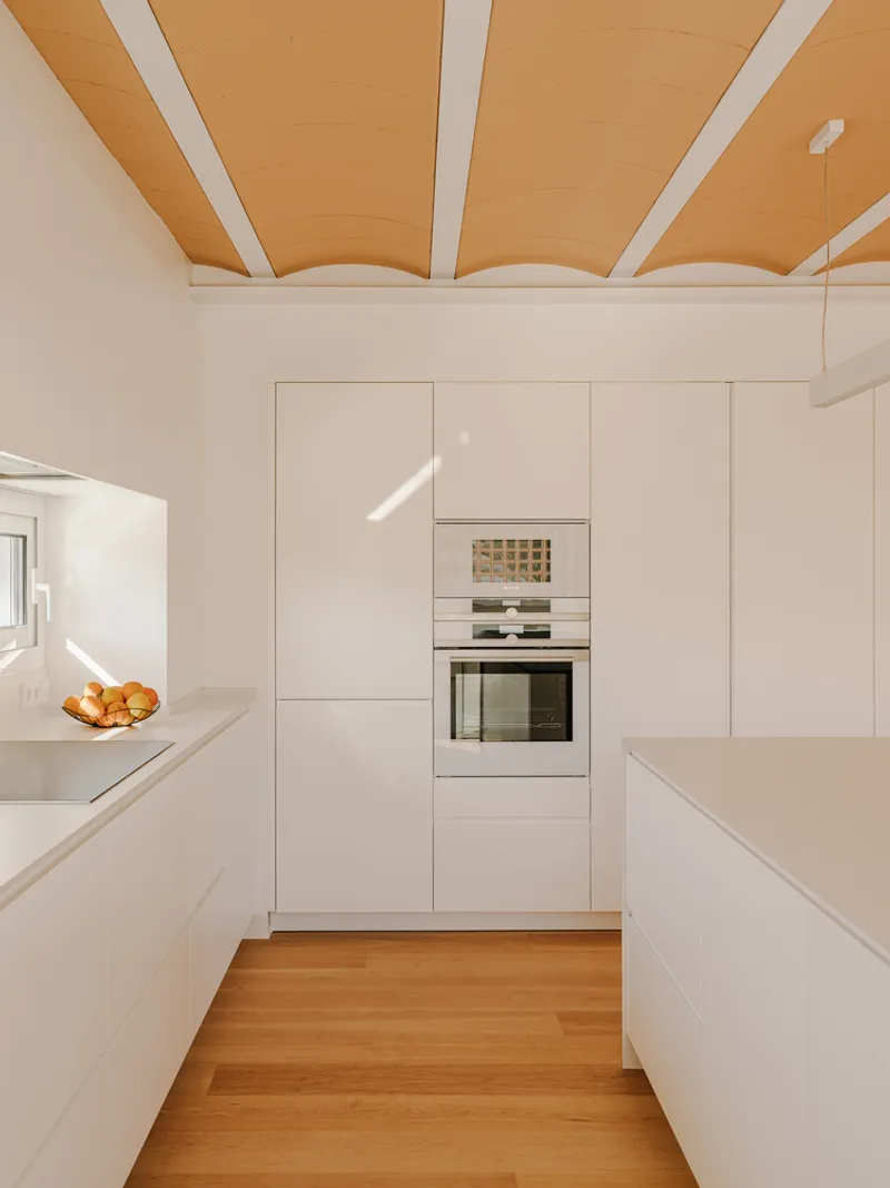 White galley kitchen with built-in appliances and timber flooring under plywood ceiling with white beams