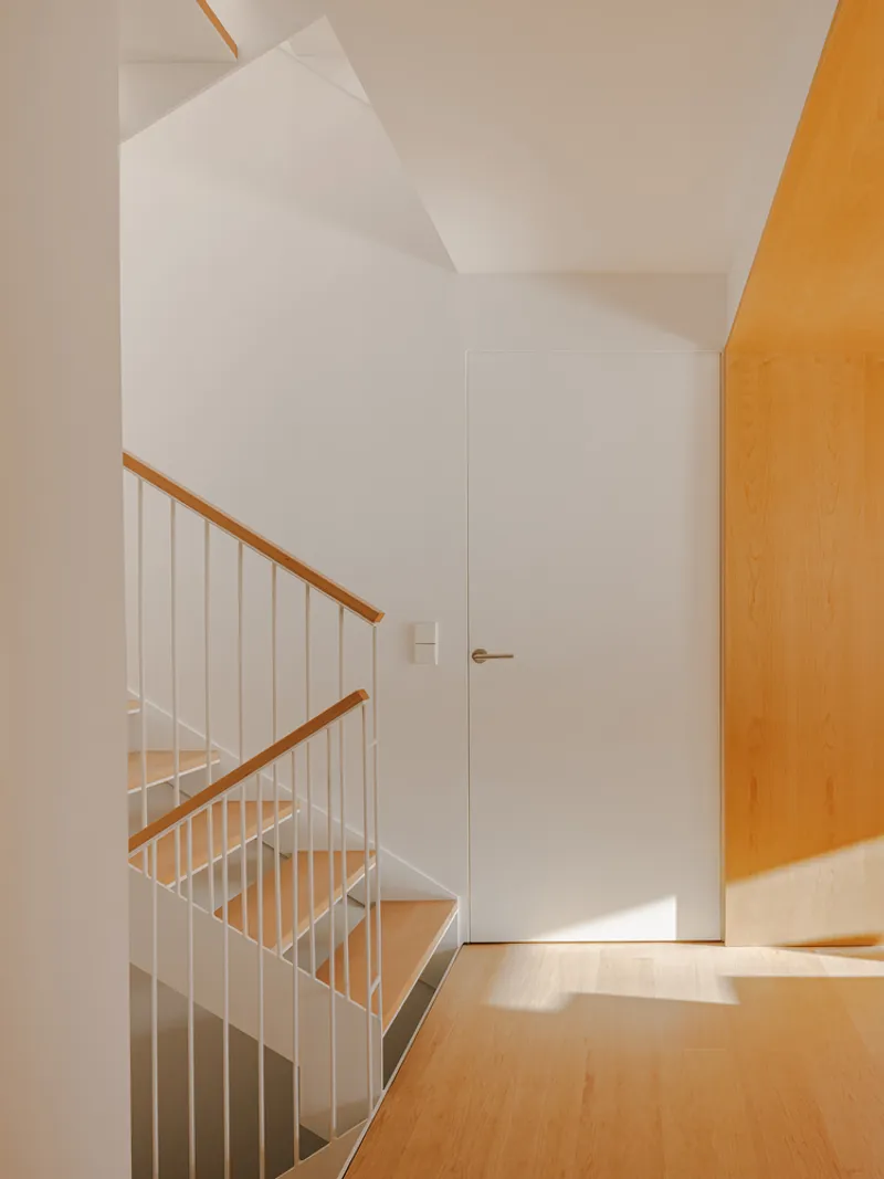 Staircase with white vertical balusters and timber handrail beneath white walls and sloped ceiling in sunlight