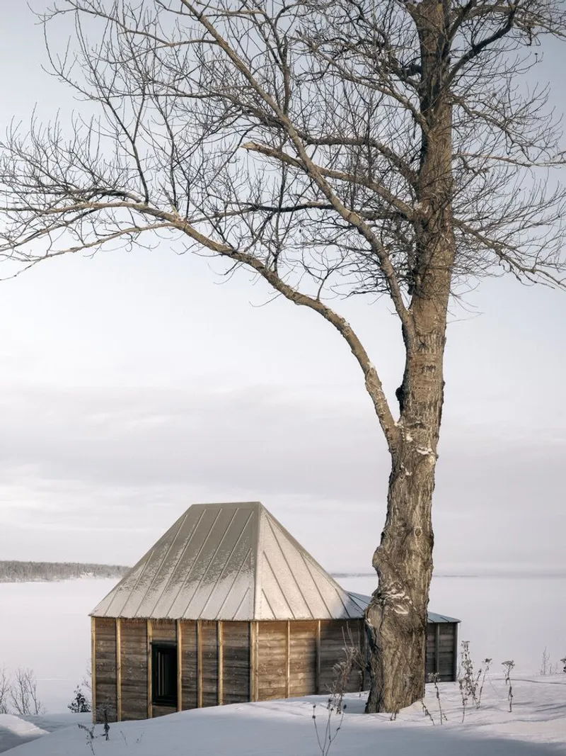 Timber-clad cabin with standing seam metal roof beside a bare tree in a snowy landscape