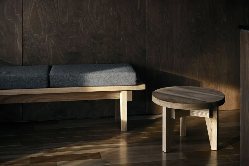Low timber bench with cushions beside a round stool against paneled wood walls in soft light