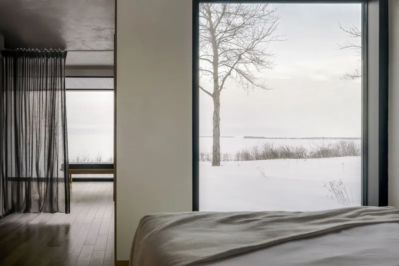 Bedroom with floor-to-ceiling glazing and sheer curtain opening to a snowy lakeshore landscape