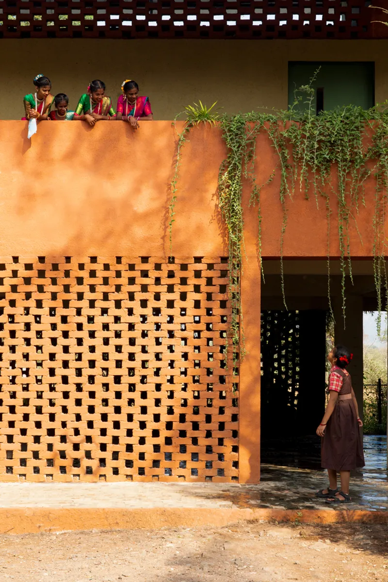 Perforated orange brick screen below a planted terrace with children looking down and figures passing through