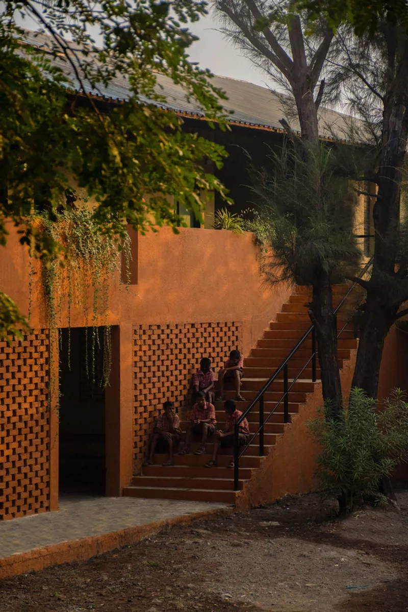 Orange staircase with perforated brick screen at dusk, with figures seated on steps beneath overhanging trees