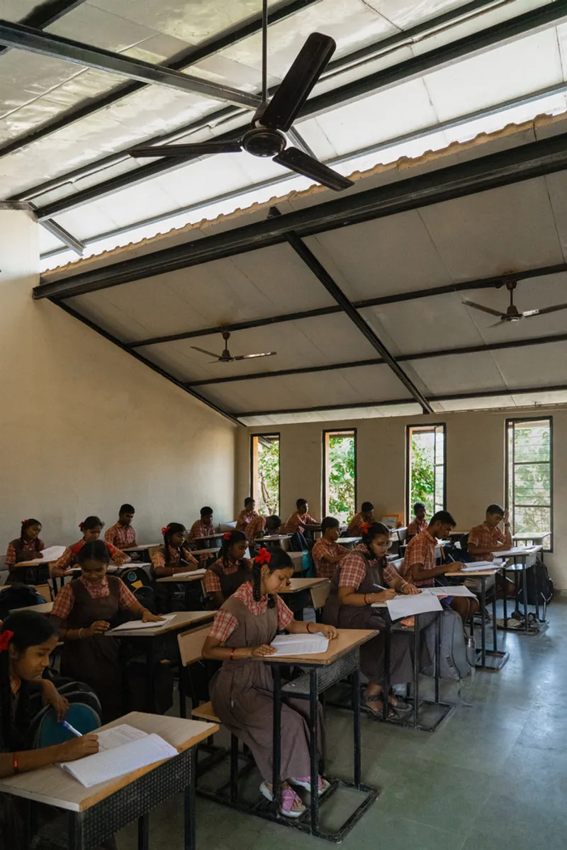 Classroom interior with exposed steel beams, translucent roof panels, ceiling fans and rows of students at wooden desks