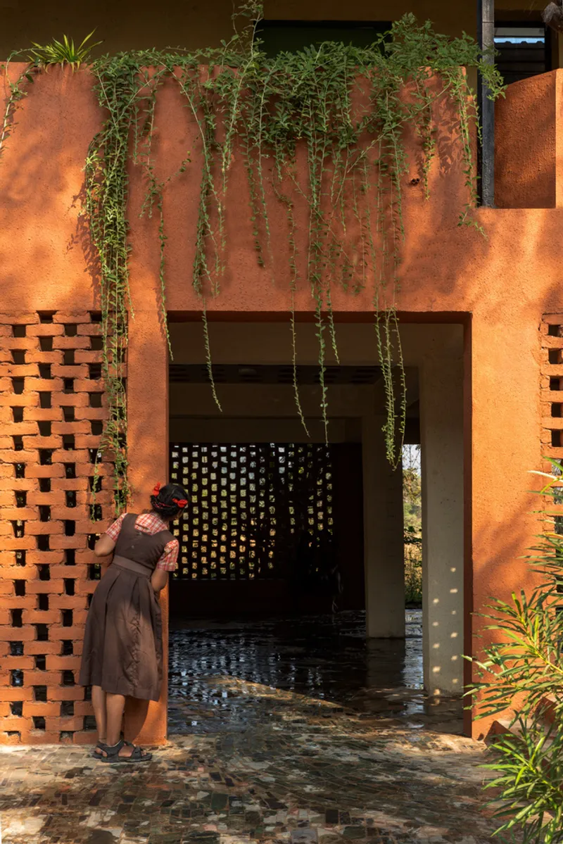 Orange entrance portal with stacked brick screen walls, trailing plants above and a figure entering dappled sunlight
