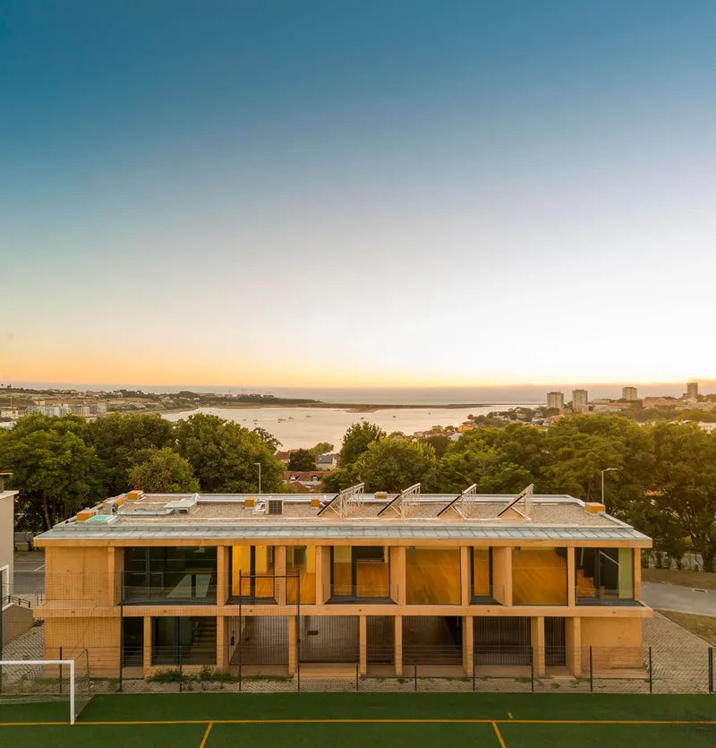 Two-story pavilion with timber louvers and sawtooth roof facing sports pitch at dusk