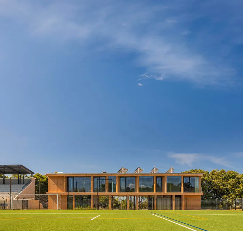 Two-story timber-clad pavilion with sawtooth roof monitors overlooking a sports field under blue sky
