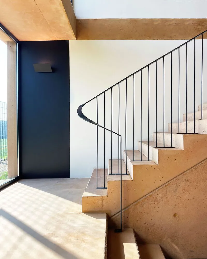 Curving staircase with slender metal railings and rammed earth steps lit by morning sunlight