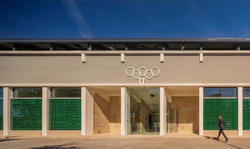 Entrance facade with green tile flanking panels and a person walking beneath an Olympic rings emblem