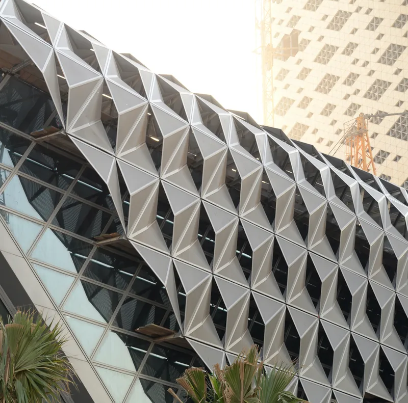 Triangulated metal canopy at ground level with palm plants beneath the diagrid tower facade