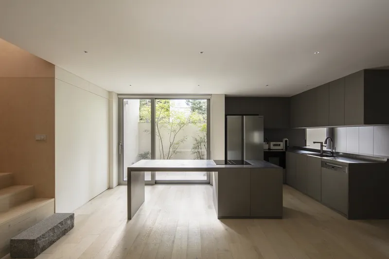 Open kitchen with grey cabinetry and island overlooking a courtyard with a young tree through floor-to-ceiling glazing