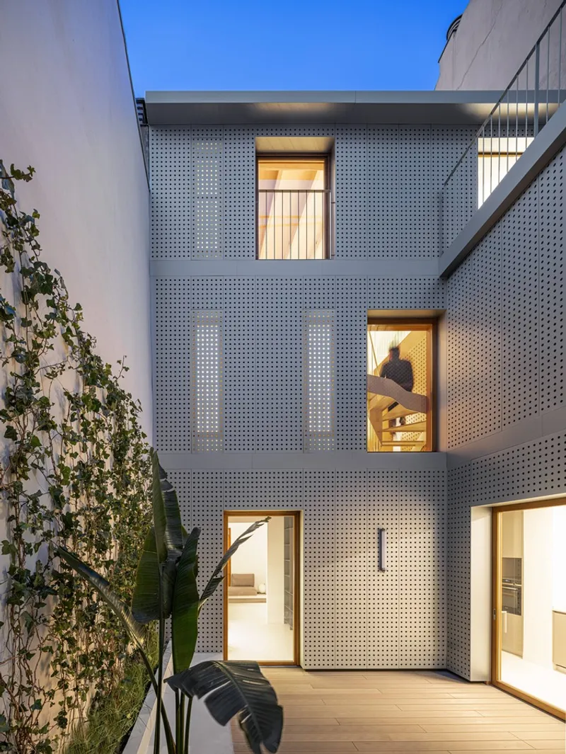 Perforated metal facade illuminated at dusk with climbing plants along the courtyard wall