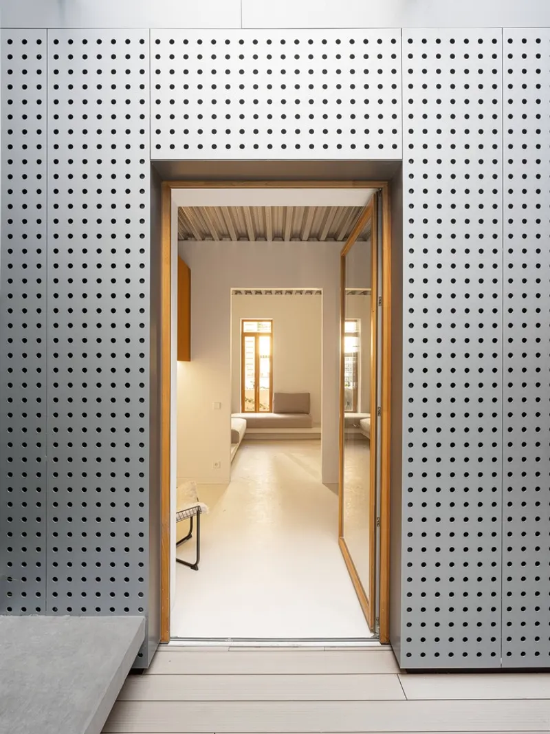 Doorway through perforated metal panels revealing an interior corridor with slatted timber ceiling