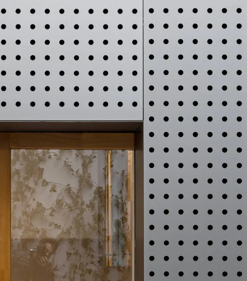 Perforated metal facade panel meeting a timber doorframe with floral glazing visible beyond