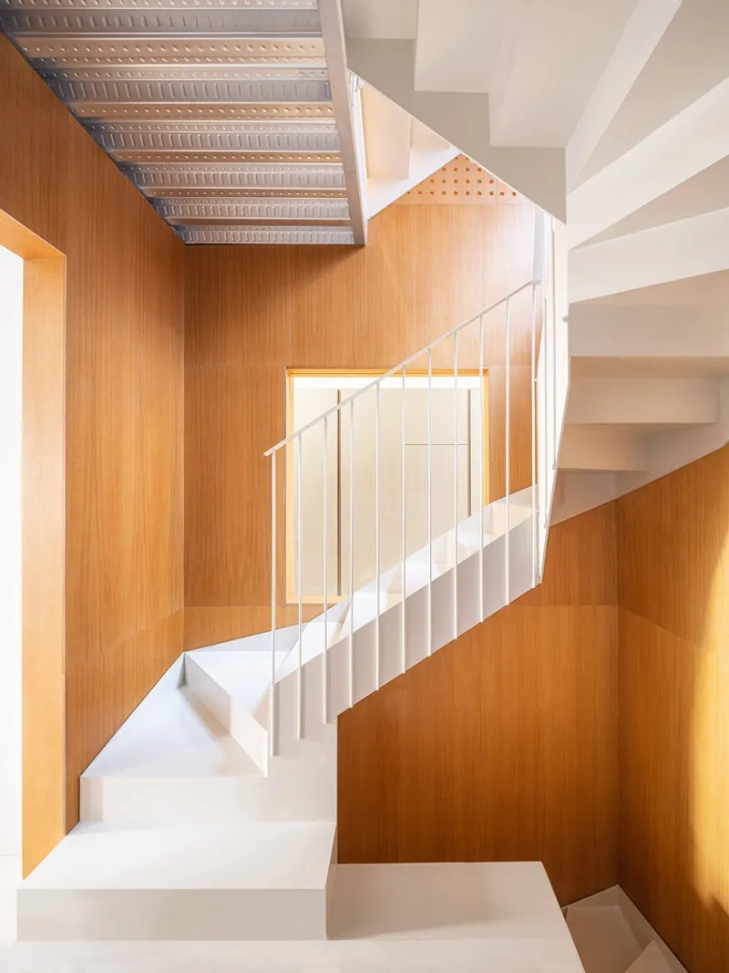 White steel staircase rising through a timber-lined shaft with perforated metal ceiling panels above
