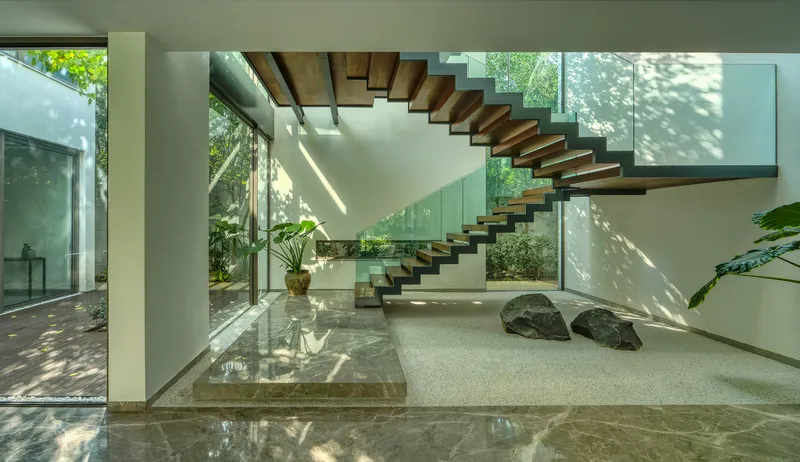 Floating timber staircase with glass balustrade ascending through a double-height courtyard with dappled sunlight