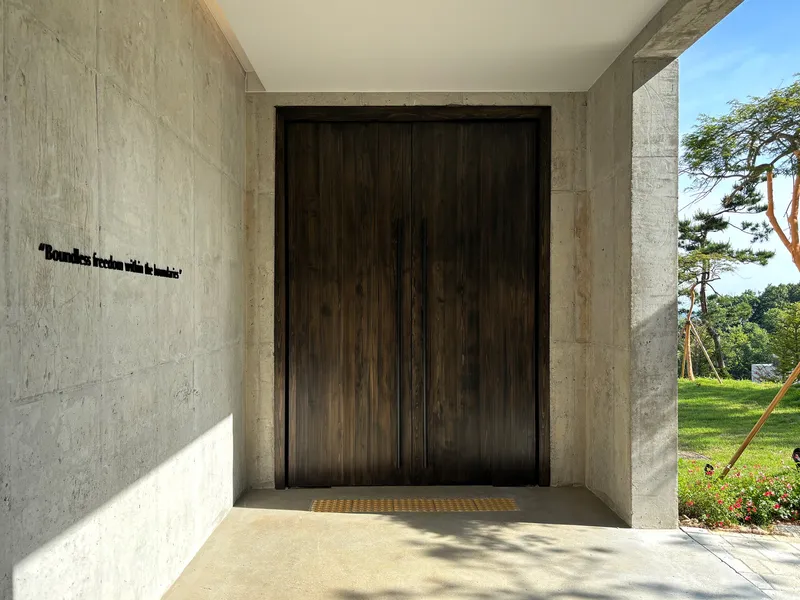 Recessed entrance with dark timber door set in concrete frame, brass mat and garden beyond