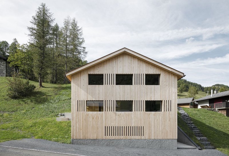 Vertical timber-clad gable facade with rhythmic window openings and wooden slat screens on a grassy hillside