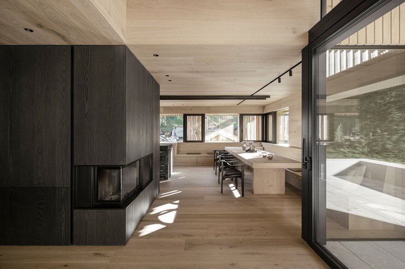 Open-plan living area with dark timber fireplace, pale wood ceiling, and full-height glazing in afternoon light