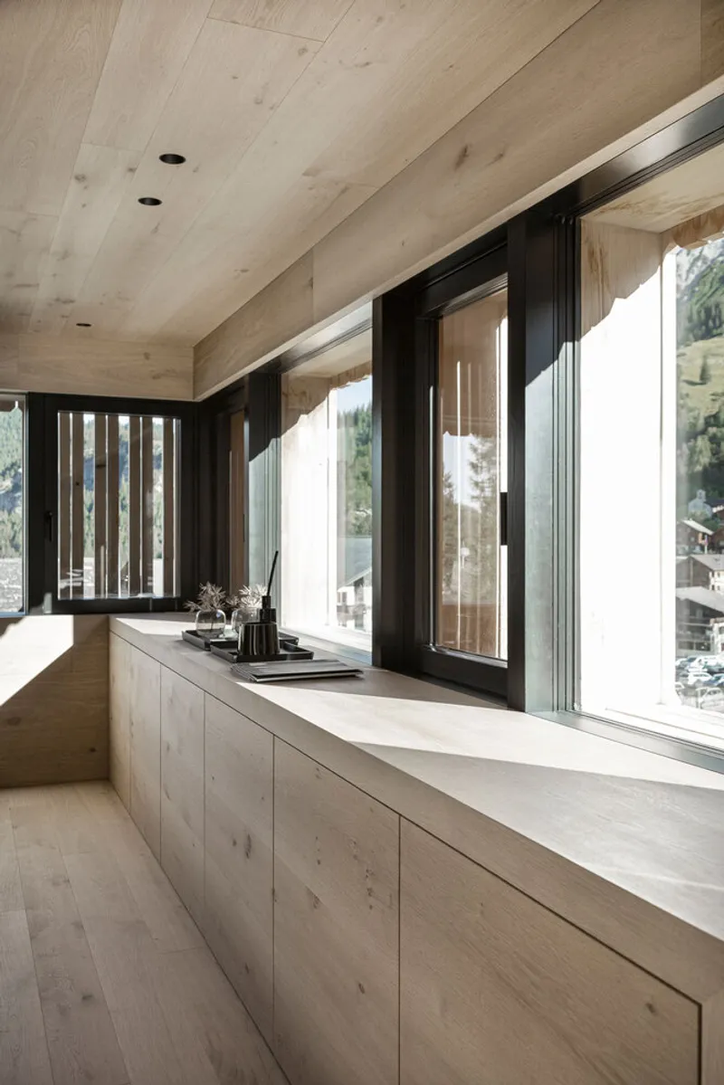 Pale timber countertop and cabinetry along a row of black-framed windows overlooking a hillside village