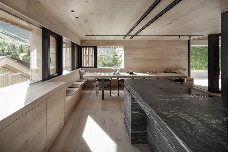 Open-plan kitchen and dining area with pale timber walls, dark stone island and sunlight streaming through corner windows