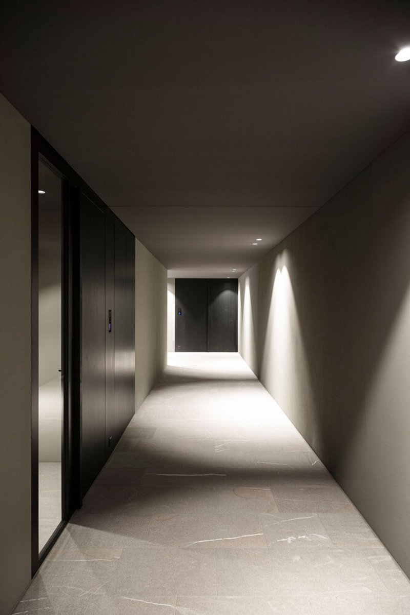 Concrete-floored corridor with darkened walls and recessed lighting leading to a backlit doorway at the end