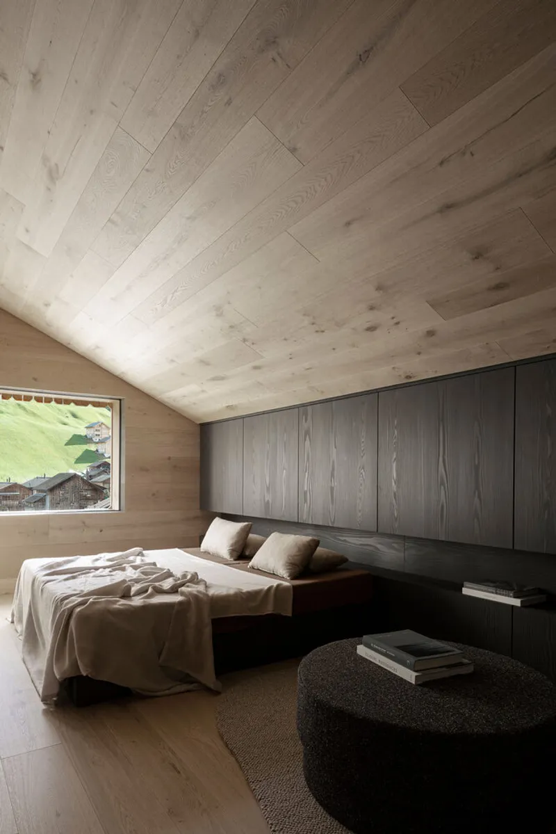 Bedroom with vaulted timber ceiling, dark wood headboard wall and picture window framing green hillside chalets