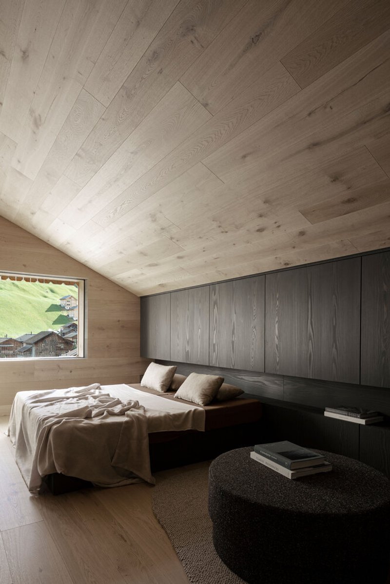 Bedroom with vaulted timber ceiling, dark wood headboard wall and picture window framing green hillside chalets