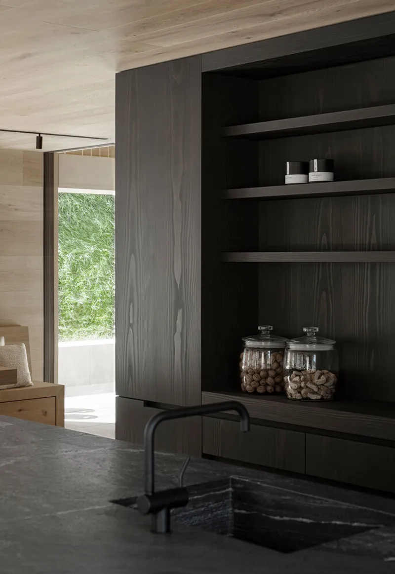 Kitchen sink view with dark timber shelving and glimpse of green courtyard through doorway