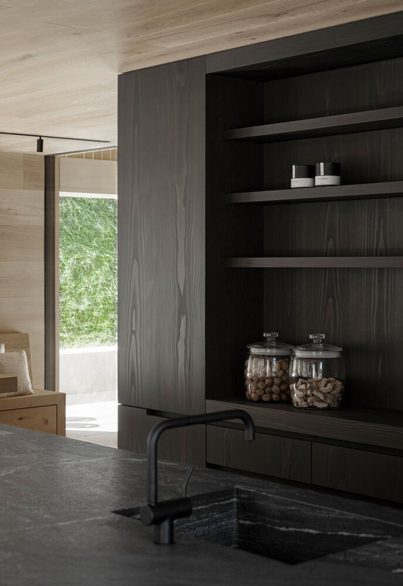 Kitchen sink view with dark timber shelving and glimpse of green courtyard through doorway