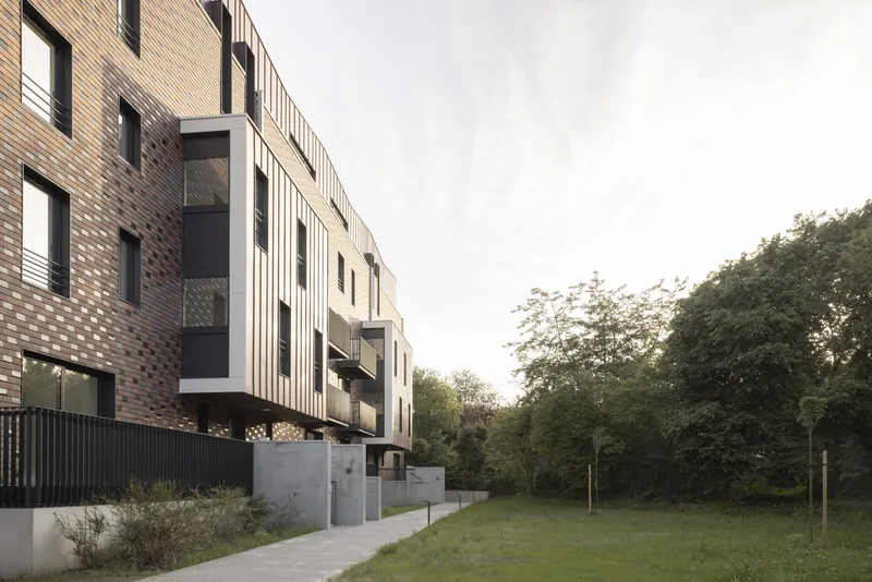 Brick facade with vertical metal panels and balconies overlooking a green lawn and trees