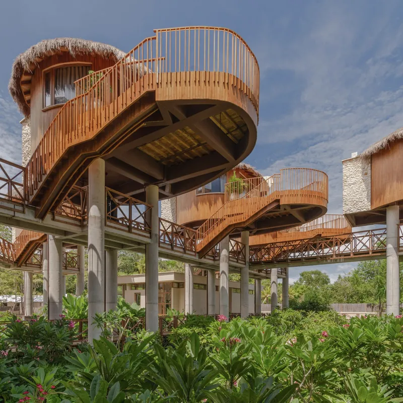 Circular elevated units with thatched roofs and vertical timber screens on concrete pilotis above flowering shrubs