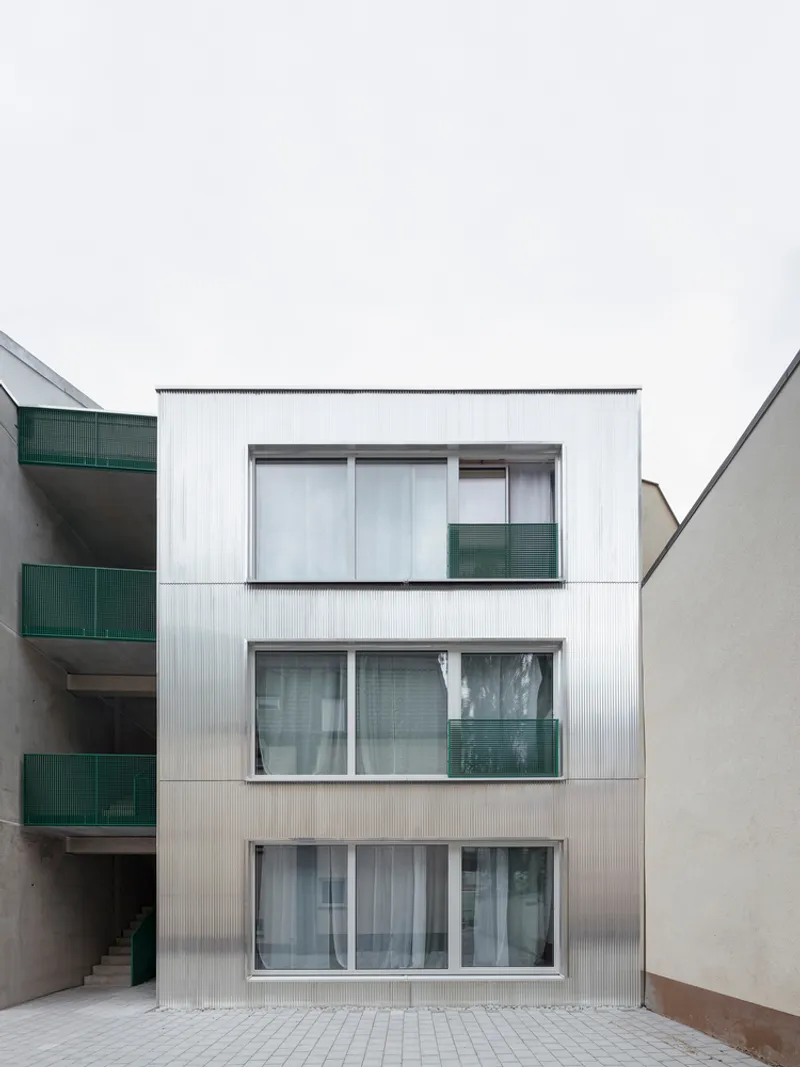 Three-story street facade with vertical corrugated cladding, sliding glass doors, and green balcony panels