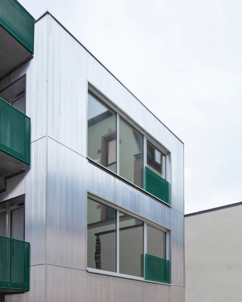 Angled view of corrugated metal facade with horizontal window bands and green balcony screens