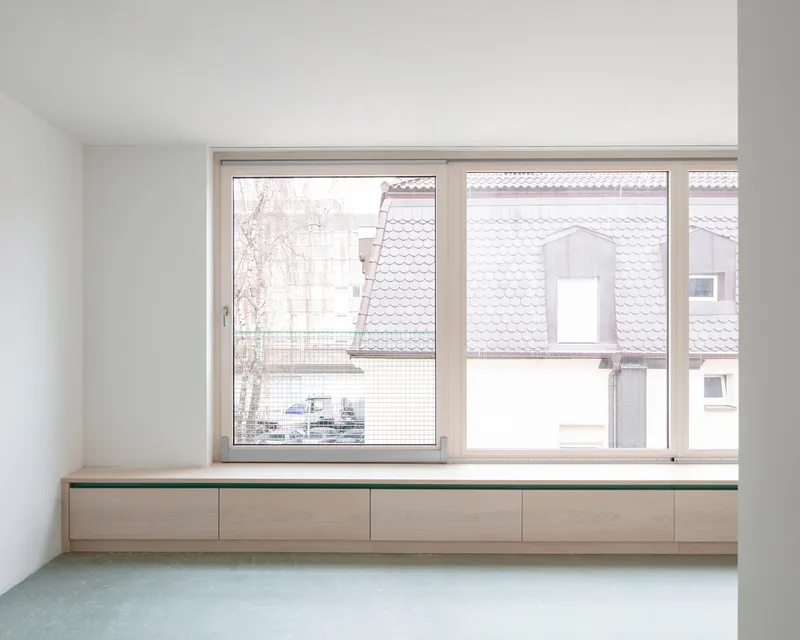 Built-in timber bench with green top below a window overlooking tiled rooftops in winter light