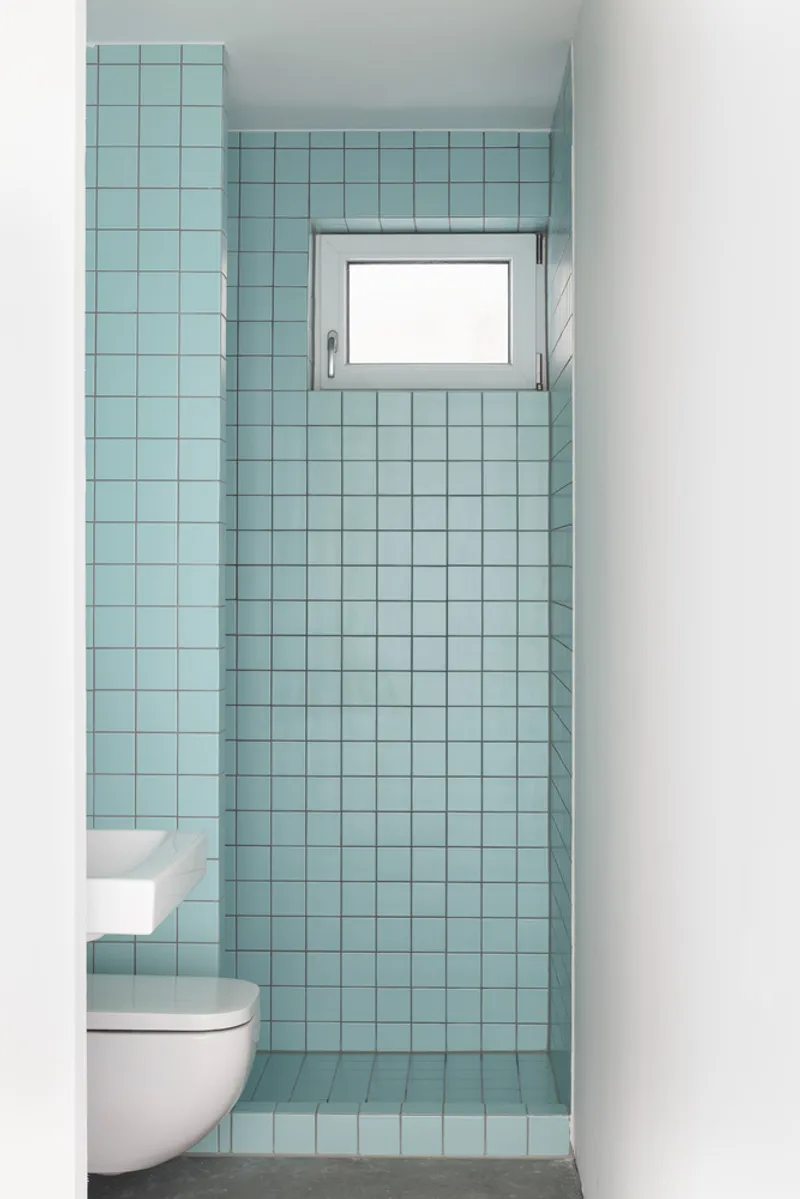 Compact bathroom with pale blue square tiles surrounding a wall-mounted toilet and high window
