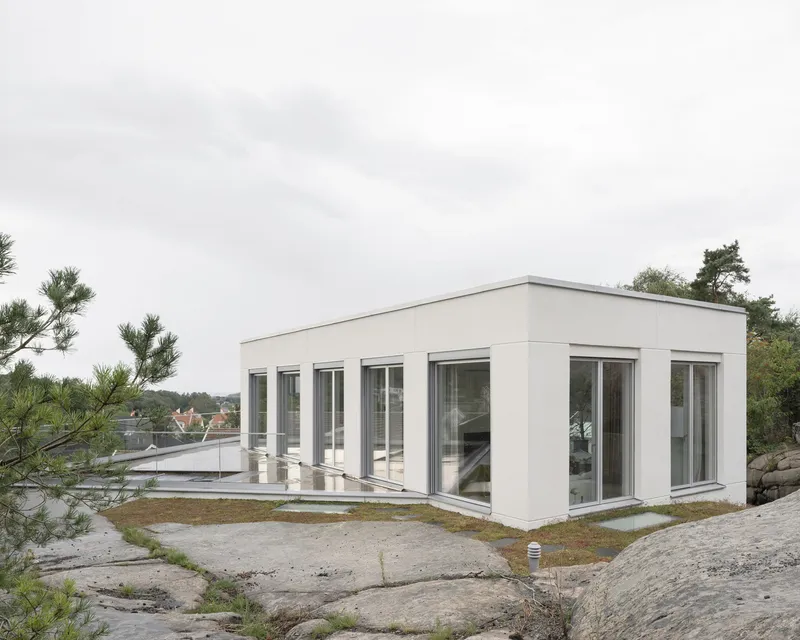 Flat-roofed pavilion with floor-to-ceiling glazing on three sides sitting on exposed bedrock under overcast skies