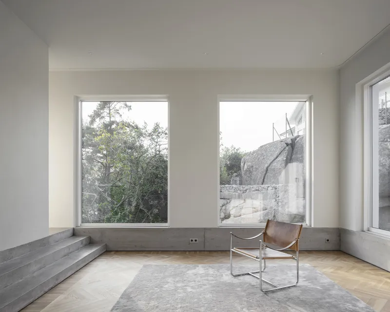 Interior room with herringbone oak floor, concrete stepped seating, and two windows framing trees and rockface