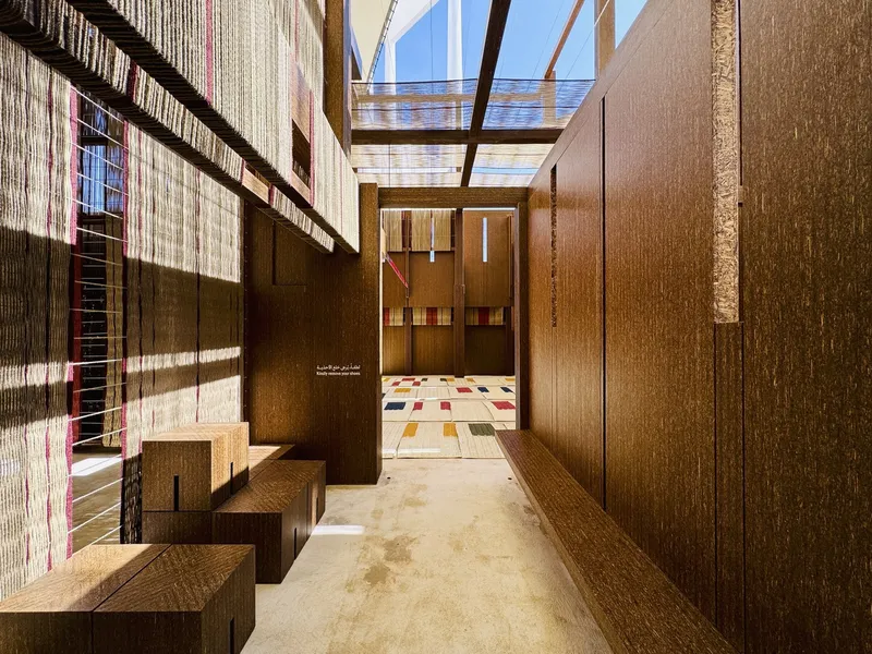 Narrow corridor with timber benches and woven screen wall casting shadow patterns toward skylit space beyond