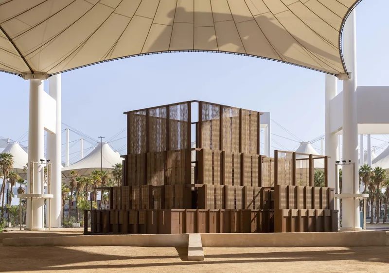 Layered timber screen pavilion beneath a tensile fabric canopy with white columns and palm trees visible beyond