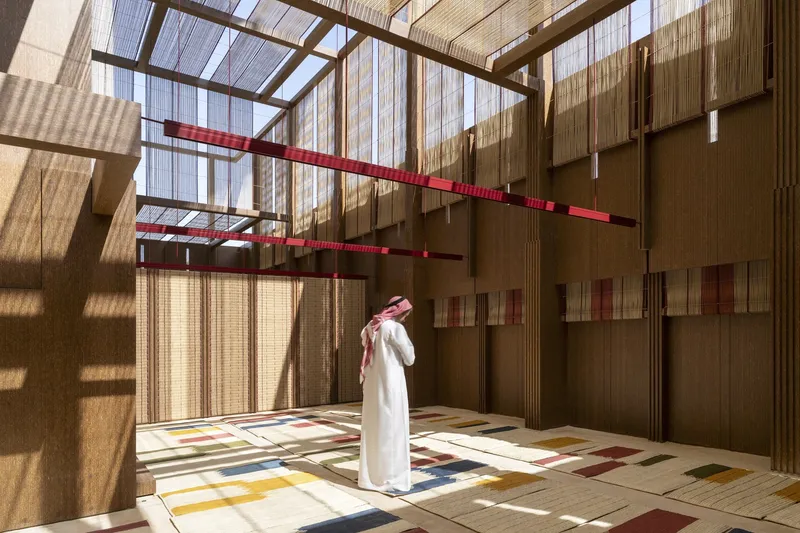 Person in traditional dress standing in sunlit interior with patterned floor tiles and suspended red fabric elements